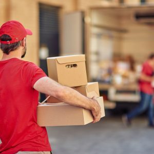 Rear view of a male courier in red uniform holding parcels for delivery while standing at storehouse, selective focus. Delivery services concept
